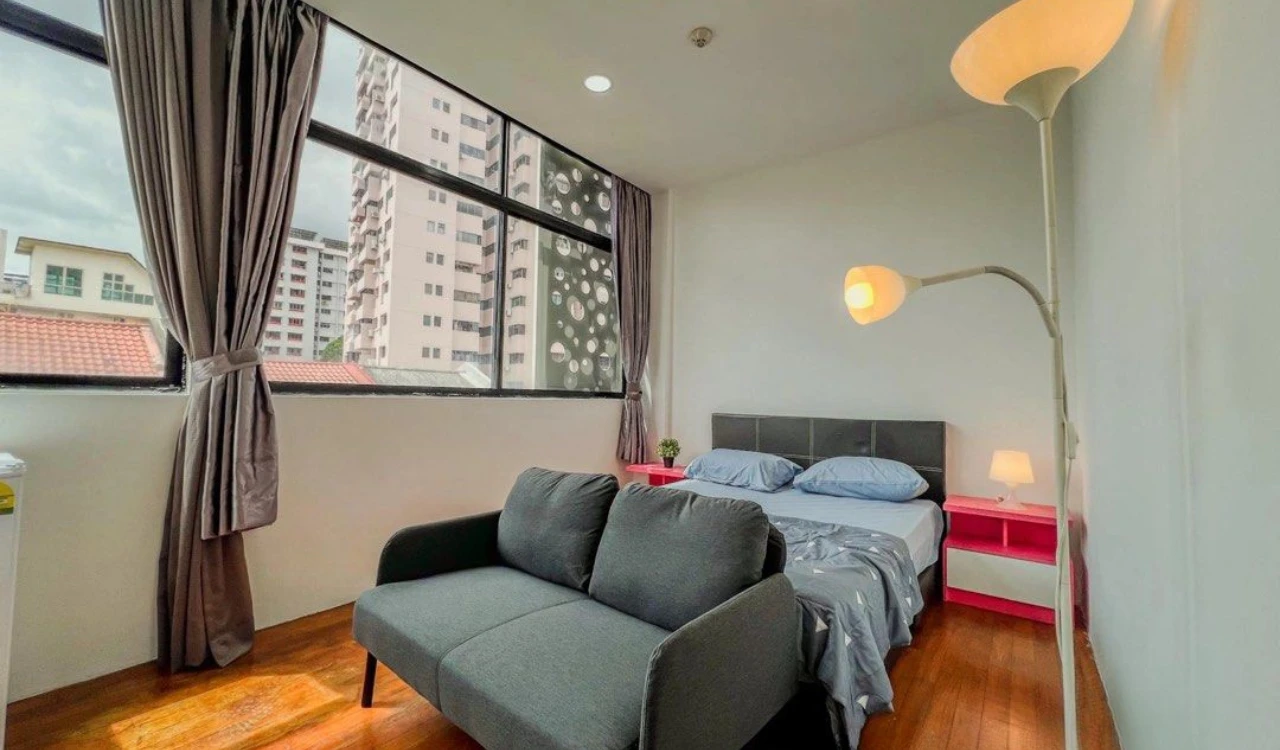 Studio apartment interior in Singapore featuring a cozy living area with a gray sofa, a bed with blue bedding, large windows, and modern lighting, ideal for singles and young couples seeking independent living.